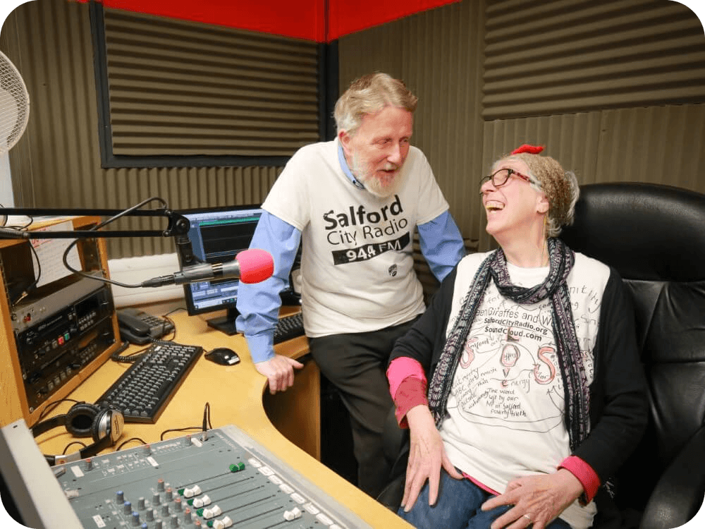 Two people smiling, sat at a microphone and radio audio desk