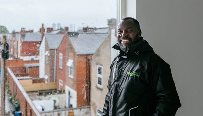 A smiling man, wearing a Salix Homes coat, standing in front of a window which overlooks the back of a row of houses