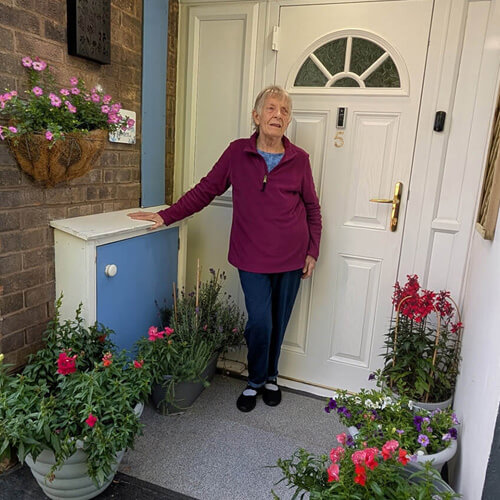 A woman standing outside the door of her apartment, surrounded by colourful pots of plants