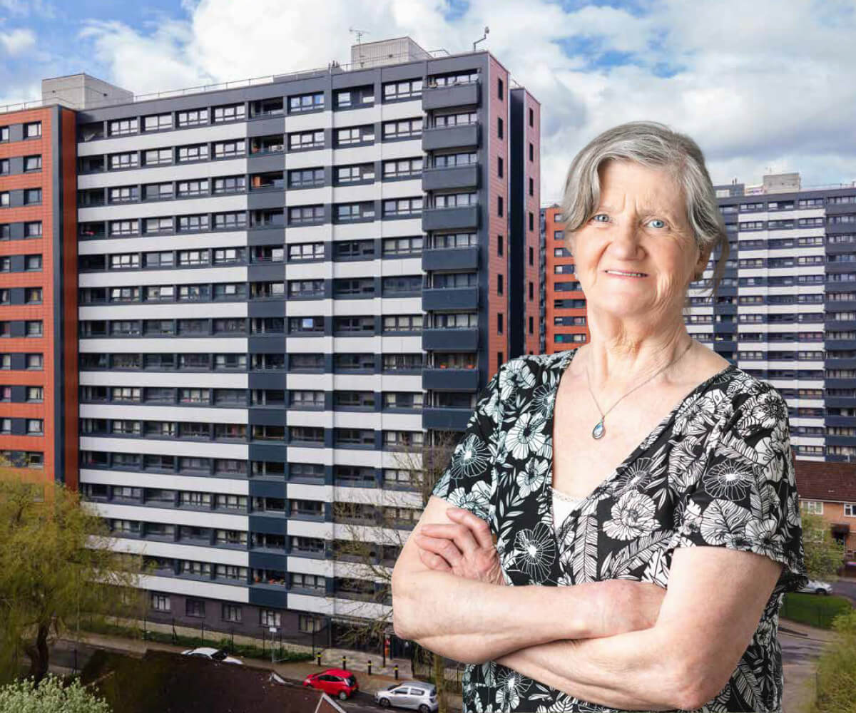 An older woman, stood with her arms folded in front of two large blocks of flats